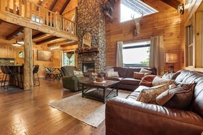 A cozy cabin interior with a stone fireplace, leather furniture, and a wooden balcony.