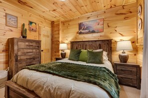 A wooden-paneled bedroom with a bed, two bedside tables with lamps, a dresser, and wall art.