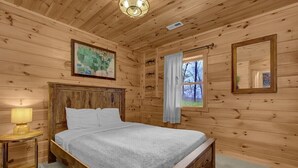 A wooden cabin bedroom with a bed, a nightstand, a map on the wall, and a window with curtains. - Arrow Ridge at Hocking Hills (Sugar Grove)