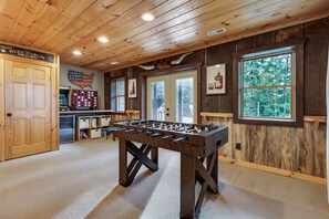 A cozy indoor space with a foosball table, wooden walls, and a large window offering a view of trees.