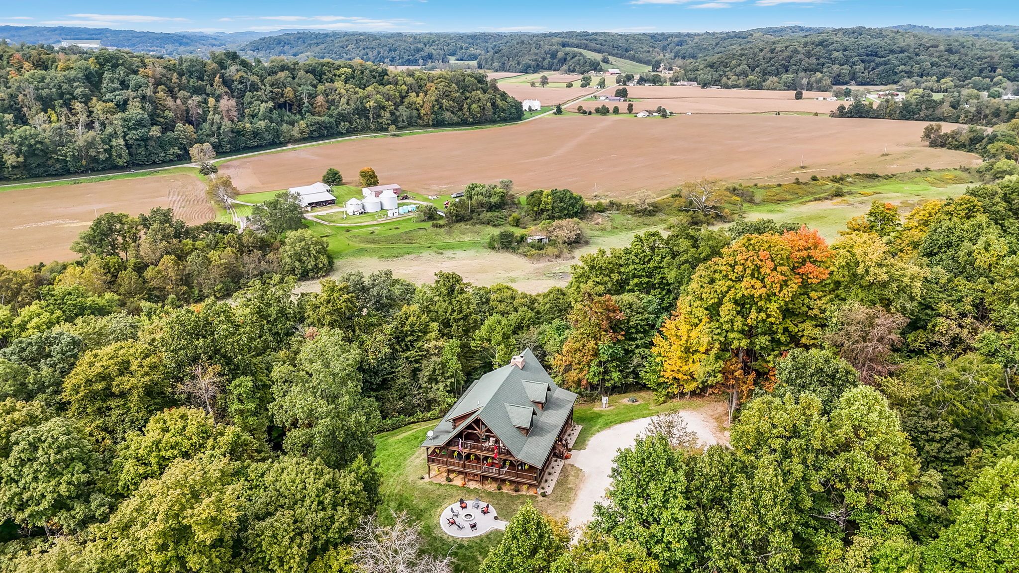 A lodge with a large porch and outdoor seating area, surrounded by dense forest and open fields.