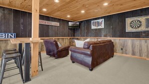 A room with a wooden ceiling, a dartboard, a couch, and a bar area with stools. - Arrow Ridge at Hocking Hills (Sugar Grove)