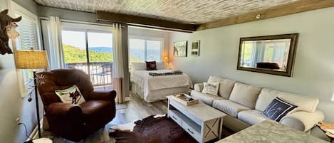 A room with a bed, a sofa, a chair, and a dining table. There is a large window with a view of the outdoors.