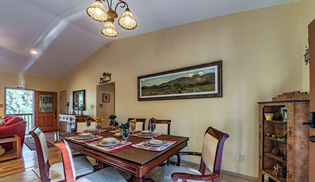A dining area with a wooden table set for six, surrounded by cushioned chairs. A chandelier hangs from the ceiling, and a framed landscape painting is on the wall.