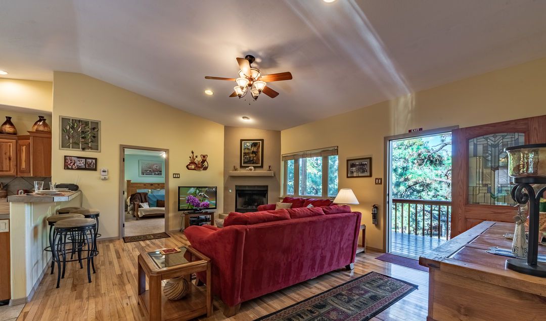 A spacious living room with a red sofa, wooden flooring, and a ceiling fan. There is a kitchen area with wooden cabinets and a dining table.