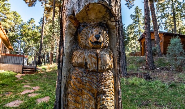 A rustic cabin with a bear sculpture carved into a tree trunk, surrounded by a forested area.