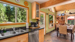 Una cocina con un amplio ventanal, armarios de madera, lavavajillas y una zona de comedor con mesa y sillas.