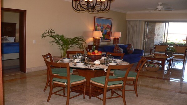 A dining area with a round table set for six, surrounded by wooden chairs with green cushions. There is a large potted plant to the left and a painting on the wall above the table. - DEC 23-DEC 30, 2024 SUPER PRESIDENTIAL SUITE 2bd/2ba private patio+jacuzzi (Cabo San Lucas)