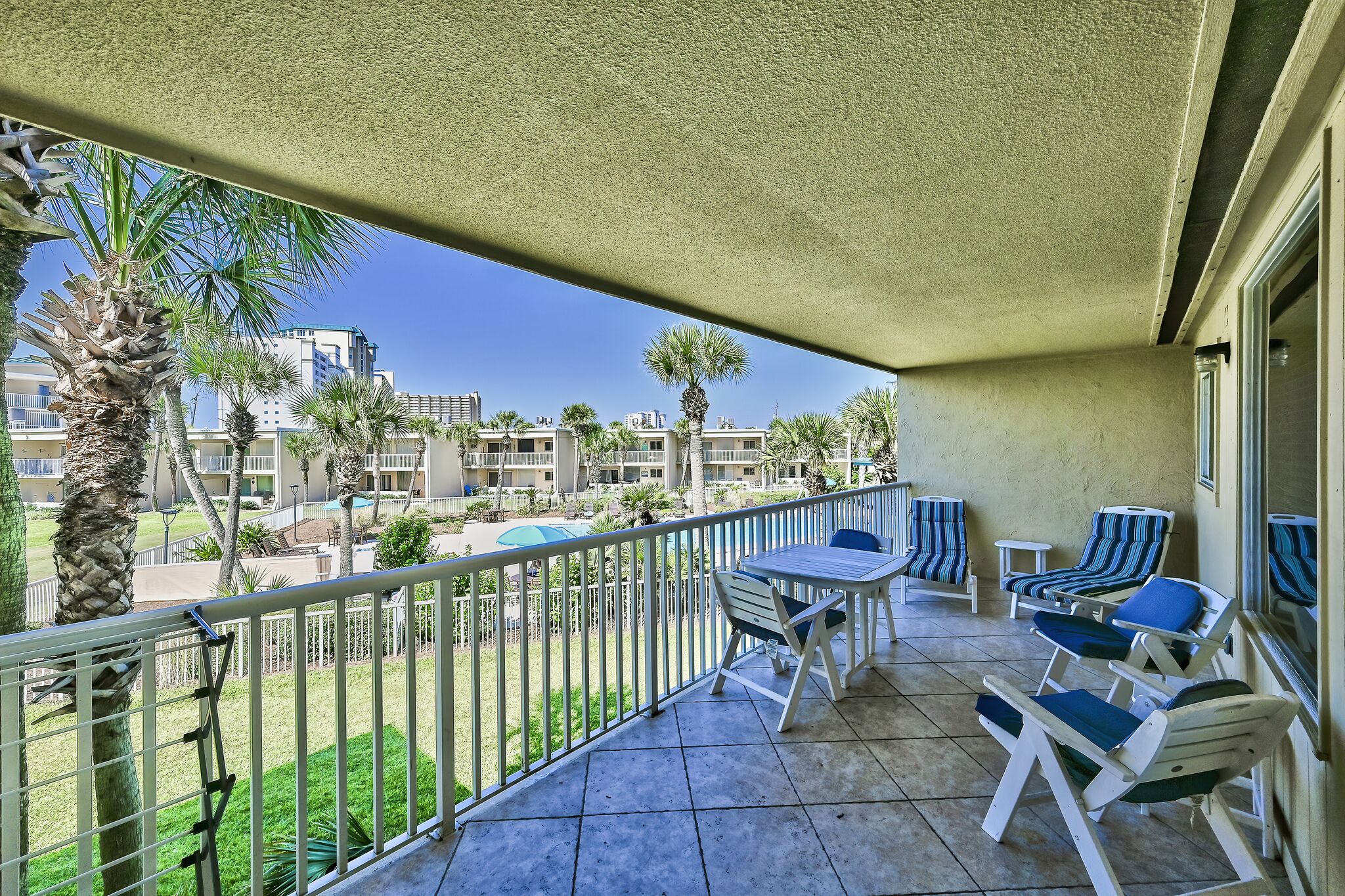 A balcony with a table and chairs, a view of a pool and buildings, and a palm tree.