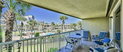A balcony with a table and chairs, a view of a pool and buildings, and a palm tree.