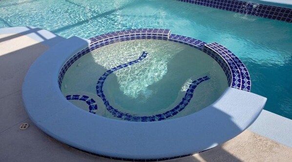 A circular hot tub with mosaic tiles, surrounded by a pool with clear blue water.
