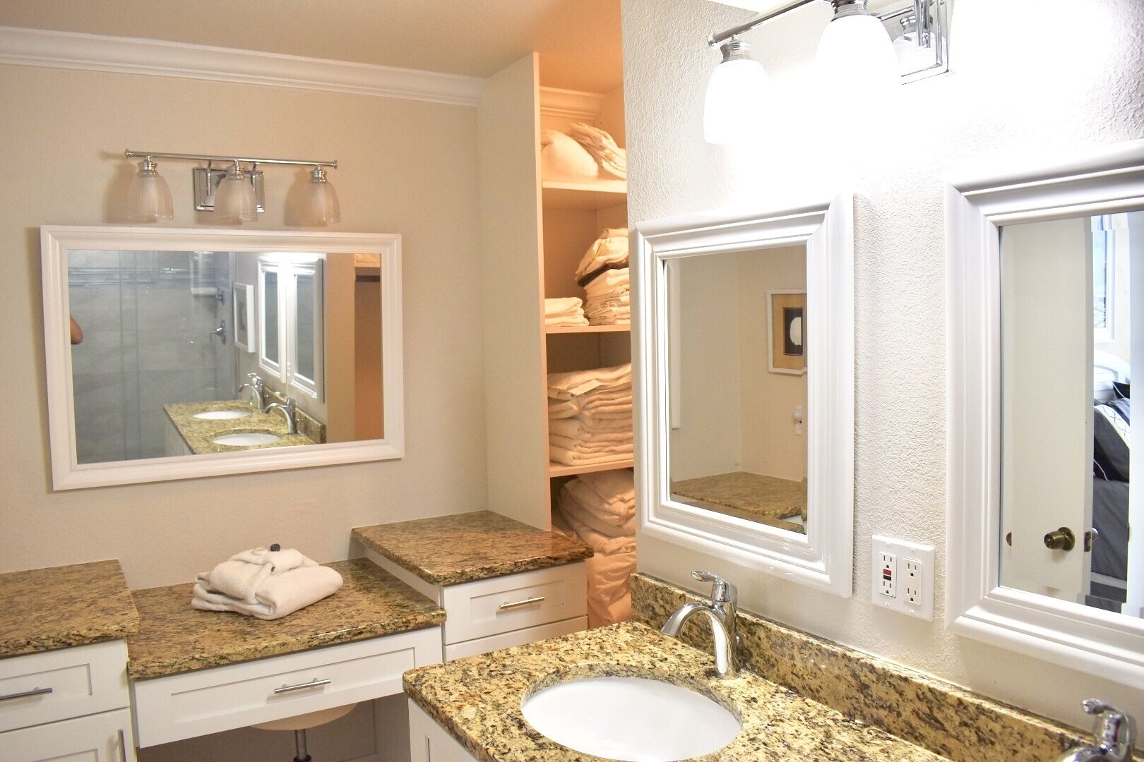 A bathroom with two sinks, a mirror, and a towel rack. There is a storage closet with folded towels.