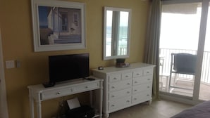 Habitación de hotel con una televisión sobre un tocador blanco, un cuadro en la pared, un espejo y un balcón con una silla.