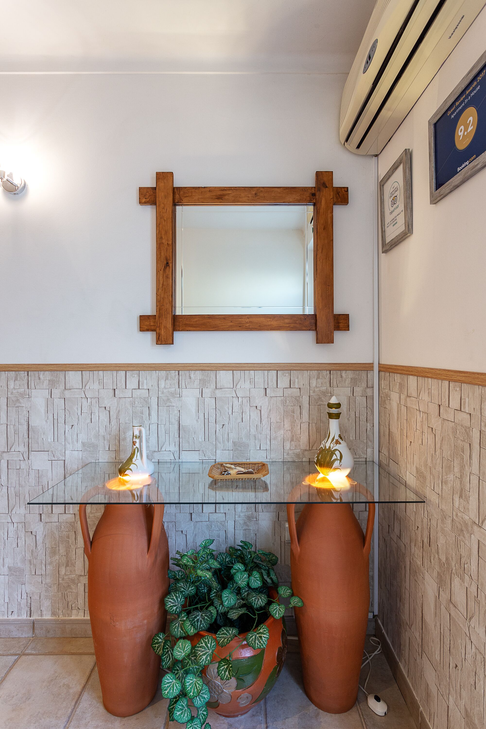 Un baño con un espejo de marco de madera, una repisa de vidrio con dos jarrones de terracota y una pared con textura.