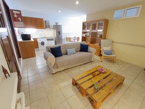 Una sala de estar con un sofá, una mesa de centro de madera y una silla. Se observa un área de cocina con armarios y electrodomésticos al fondo.