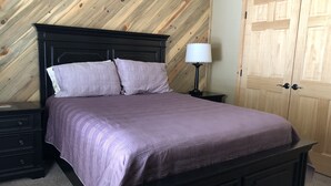 Un dormitorio con techo inclinado de madera, cabecero negro, cama con colcha morada, mesita de noche con lámpara y armario de madera.