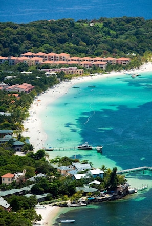 Vista aérea de um resort costeiro com vários edifícios, uma praia de areia e águas turquesa cristalinas.