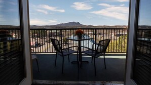 Een balkon met een tafel en stoelen, een vaas met bloemen, en uitzicht op een parkeerplaats en bergen.