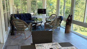 A covered patio with wicker furniture, a table with a vintage tablecloth, and a large window offering a view of greenery. - Come and enjoy a beautiful view and relaxing time at Windy Knoll.... (Afton)