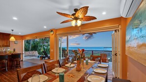 Un coin repas avec vue sur l’océan, un ventilateur de plafond et un grand tableau au mur.