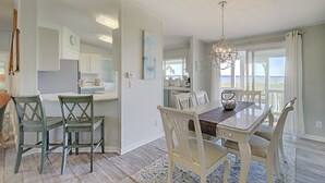 A dining area with a table and chairs, a kitchen in the background, and a view of the sea through large windows.