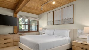 A bedroom with a large bed, wooden ceiling, a flat-screen TV, a window with blinds, and artwork on the wall.