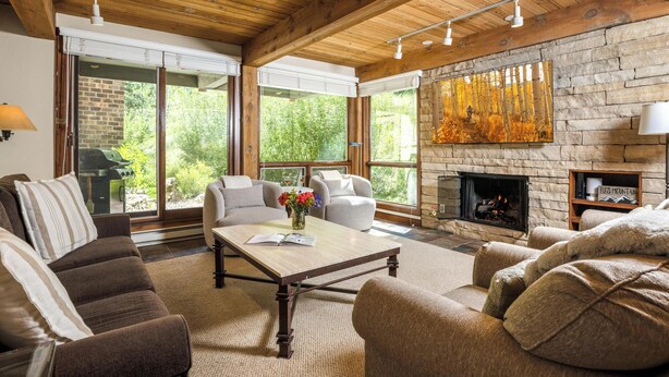 A cozy living room with a stone fireplace, large windows, and a wooden ceiling.