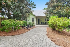A green house with a paved driveway leading to a front door, surrounded by greenery and bicycles parked outside.