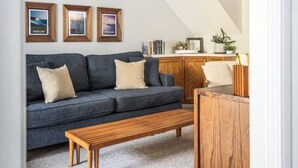 A living room with a dark blue sofa, two wooden side tables, and a wooden bench. There are three framed pictures on the wall, a kitchen area with wooden cabinets, and a staircase with a wooden railing.