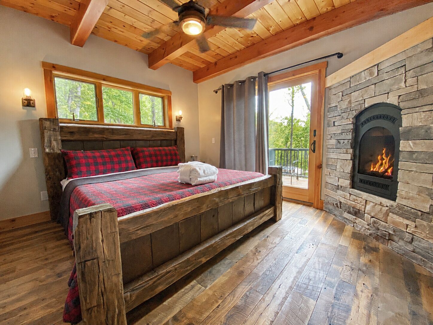 Un lit en bois recouvert d’une couverture à carreaux, une cheminée en pierre et un balcon avec des rideaux.