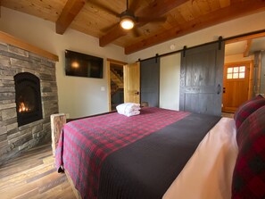 A bedroom with a stone fireplace, a bed with a red and black quilt, and a wooden ceiling. - Rustic Timber | 5-Bedroom Pet Friendly Black Mountain Retreat with Views (Fairview)