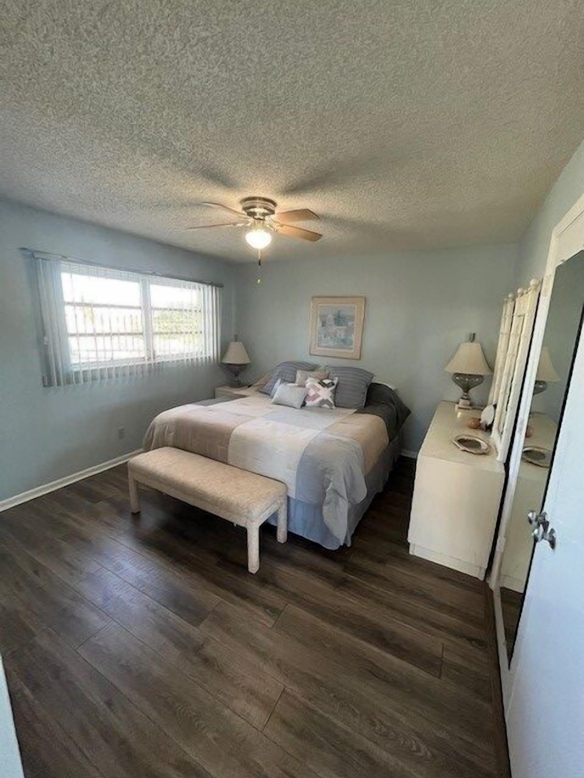 Un dormitorio con cama, banca, ventilador de techo, ventana y cómoda.