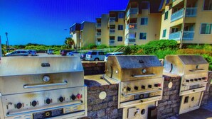 Outdoor barbecue grills, a stone wall, and a multi-story residential building with balconies. - Ocean View Condo@Beach Front Resort 150' Waterslide Indoor Pool Tennis Mini Golf (Atlantic Beach)