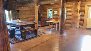 A log cabin interior with a wooden dining table, benches, a kitchen area, and a large window.