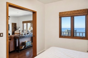 A bedroom with a bed, a window with a view of the sea, a wooden door, and a hallway leading to another room.