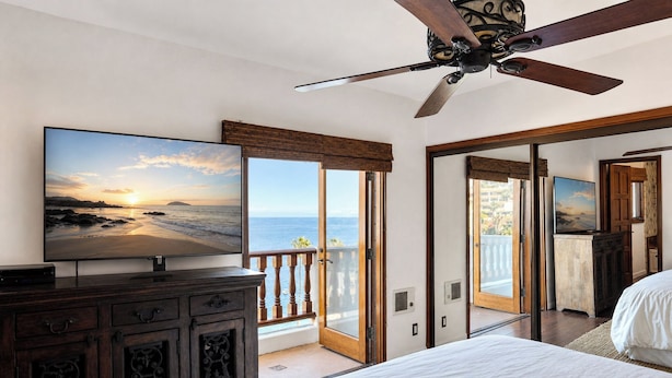 Un dormitorio con una cama grande, un ventilador de techo, un televisor sobre un mueble de madera y un balcón con vista al océano.