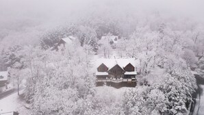 Una cabaña cubierta de nieve con techo simétrico y una chimenea central, rodeada de árboles densos.
