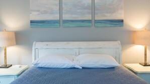 Une chambre à coucher avec un lit, une tête de lit blanche, deux lampes de chevet et trois tableaux représentant un paysage marin accrochés au mur.
