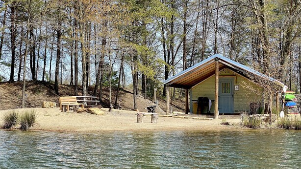 A lakeside cabin with a wooden deck, a bench, and a covered picnic table. - Castaway lake side canvas cottage scene (Jasonville)