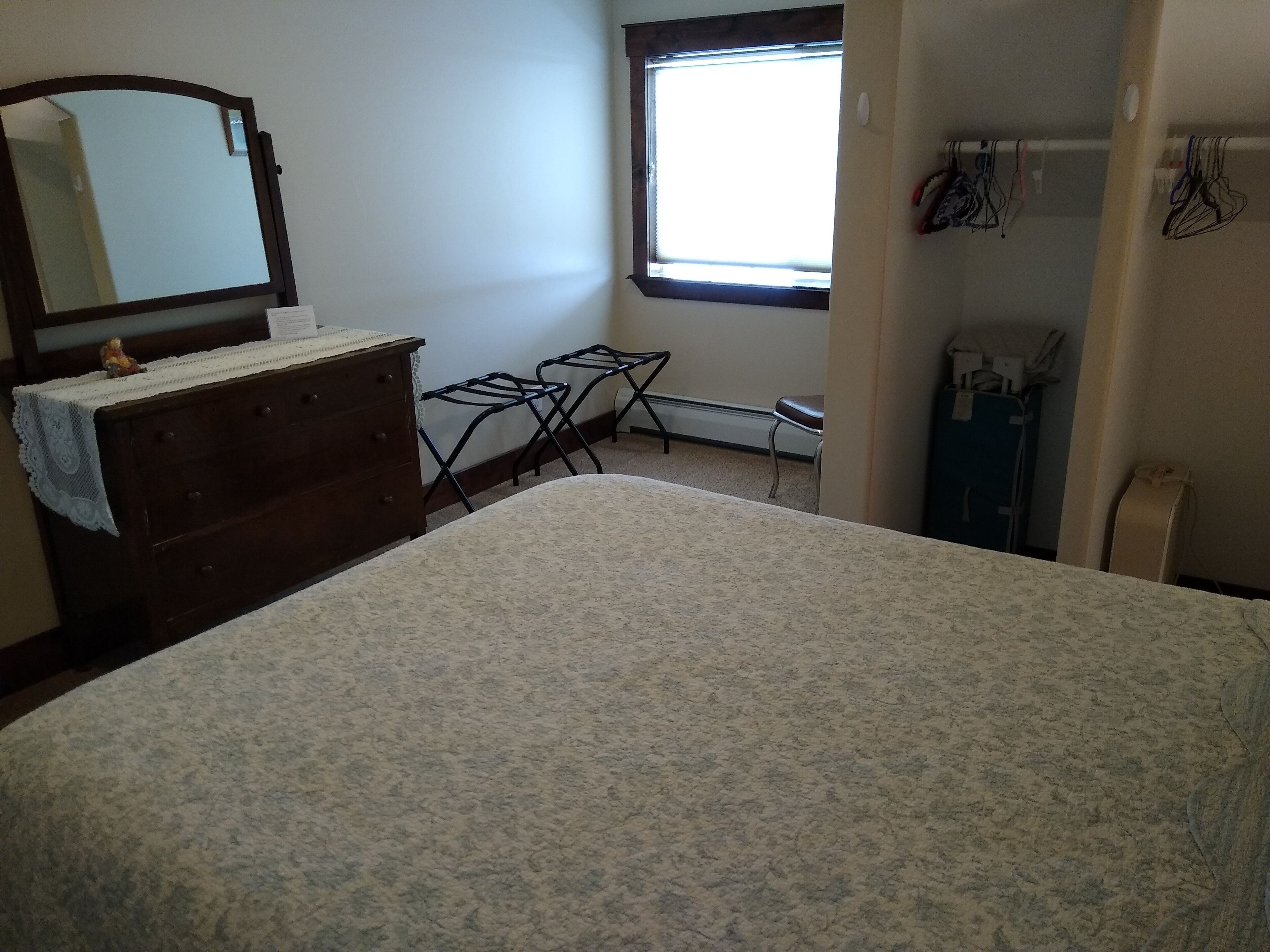 Un dormitorio con cama, cómoda, espejo, ventana y tabla de planchar.
