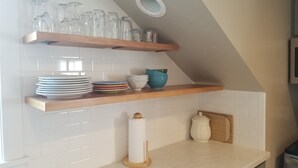 A kitchen with white tiled walls, wooden shelves, and various kitchenware including plates, bowls, and a rolling pin.
