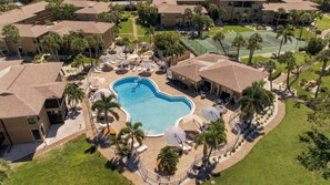 An aerial view of a residential area with a swimming pool surrounded by lounge chairs and umbrellas, and a tennis court.
