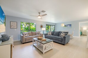 A spacious living room with two grey sofas, a white coffee table, and a ceiling fan. Large windows offer views of greenery outside.
