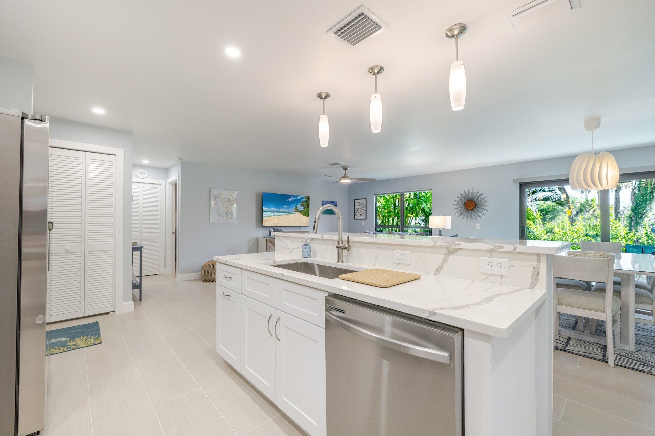 A modern kitchen with white cabinetry, a central island, stainless steel appliances, pendant lighting, and a view of greenery outside.