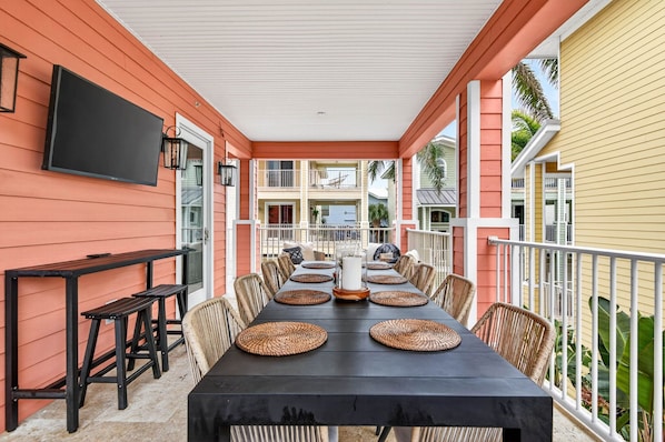 A covered outdoor dining area with a long table, chairs, and a TV mounted on the wall.