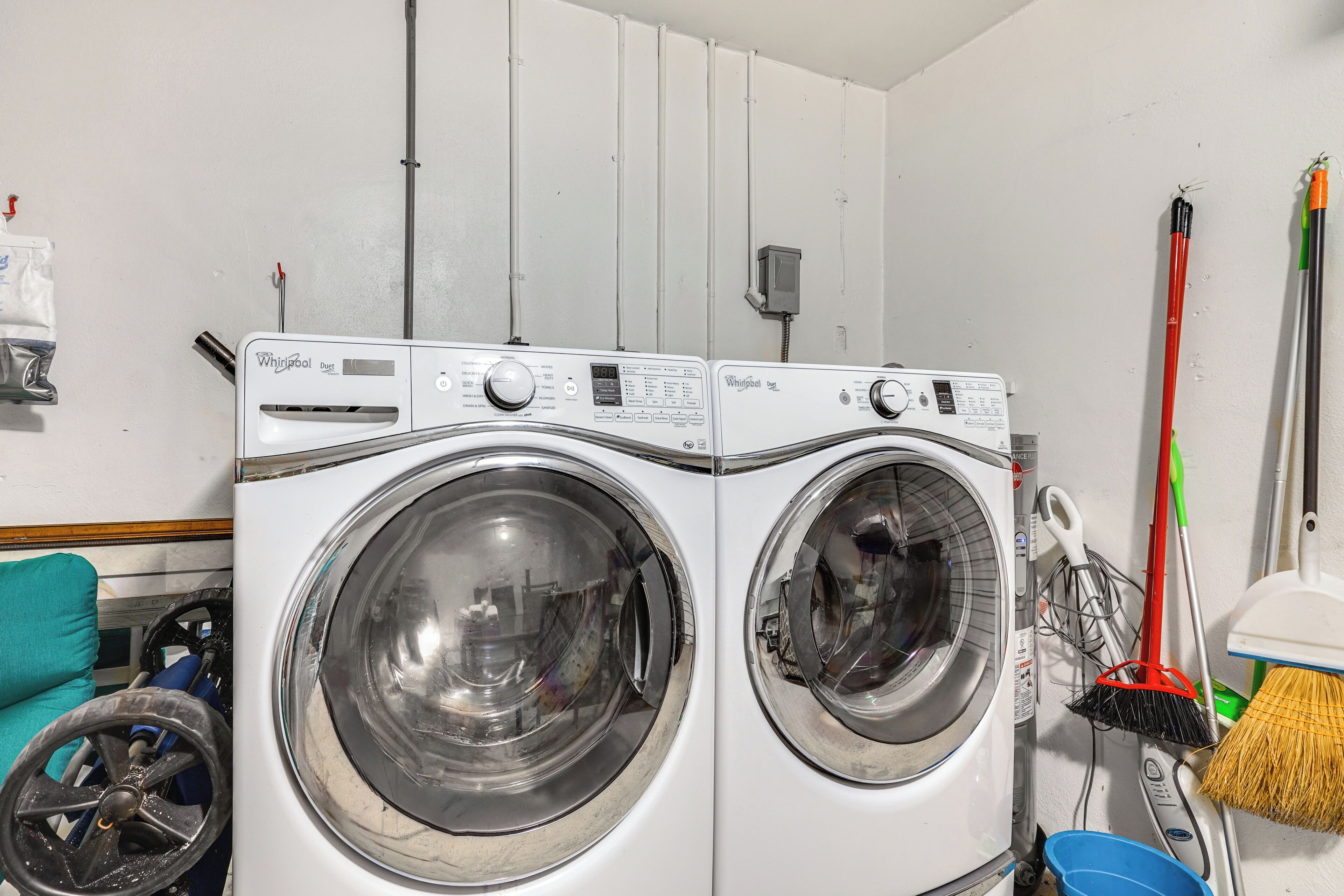 Two front-loading washing machines in a laundry room with a blue bucket and brooms.