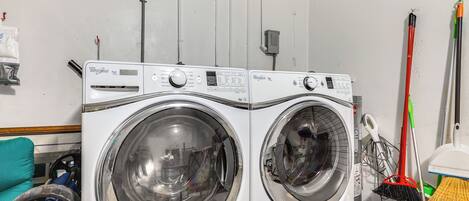 Two front-loading washing machines in a laundry room with a blue bucket and brooms.