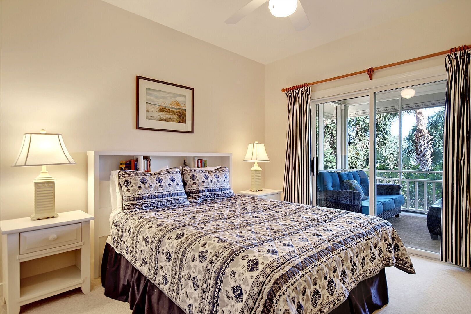 A bedroom with a large bed, two bedside lamps, a framed picture on the wall, and a balcony with a blue sofa.