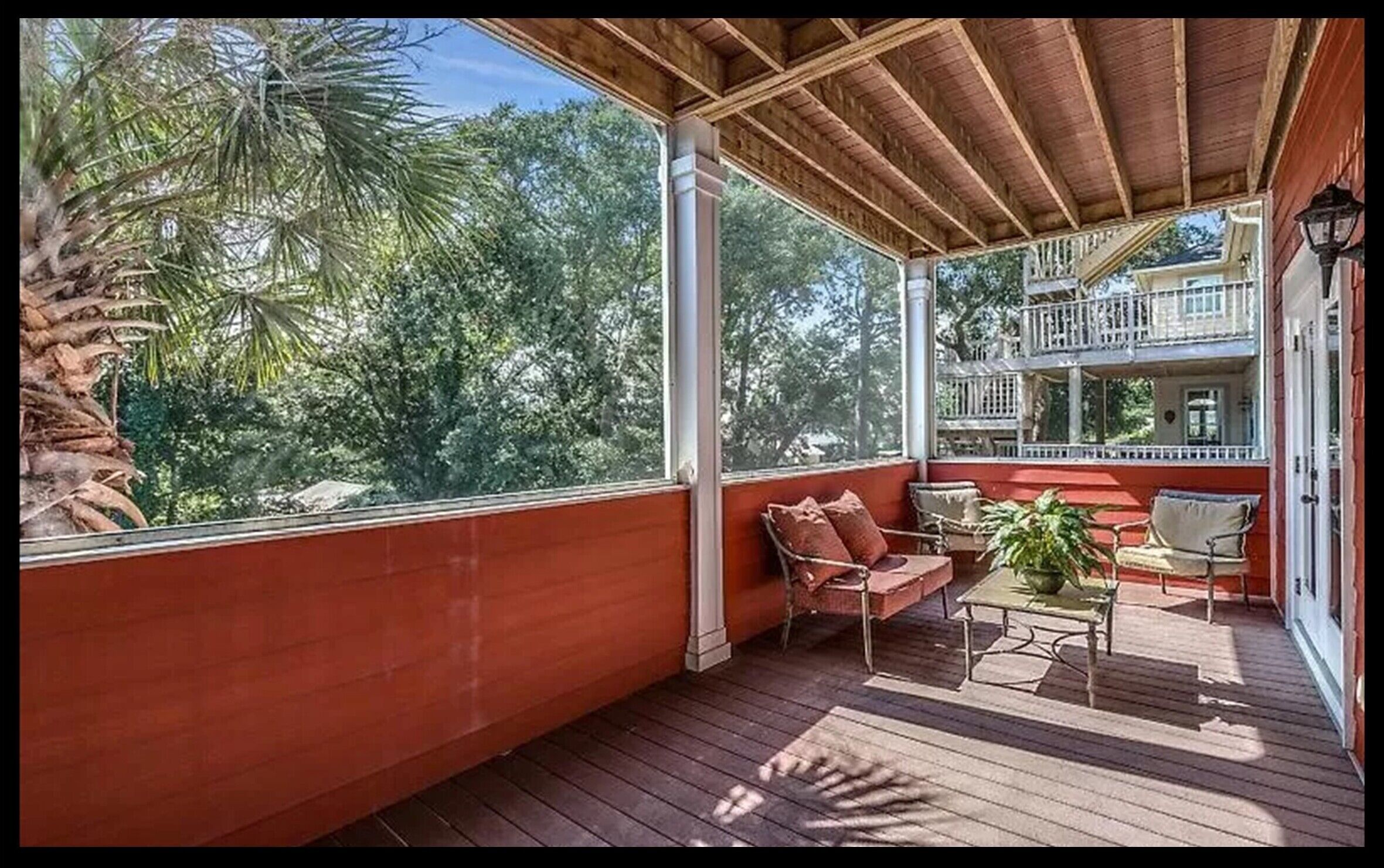 A covered outdoor area with a wooden deck, a seating area with chairs and a table, and a view of trees and a building in the background.