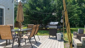 Eine Terrasse im Freien mit Grill, Esstisch, Stühlen und Sonnenschirm.
